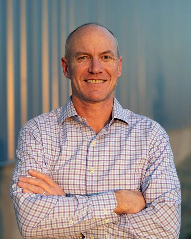 A man wearing a button-down shirt poses, smiling, with his arms folded.