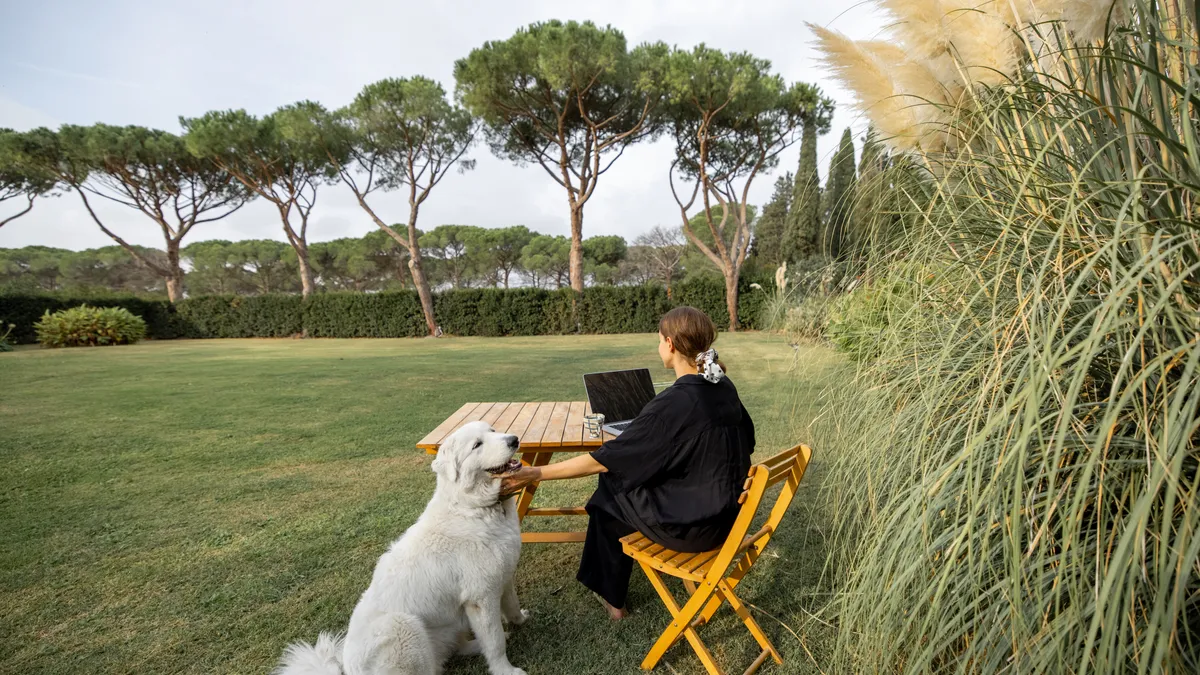 A person pets a dog as they sit at lawn table and do work on a laptop. They are in the yard of a Mediterranean villa.