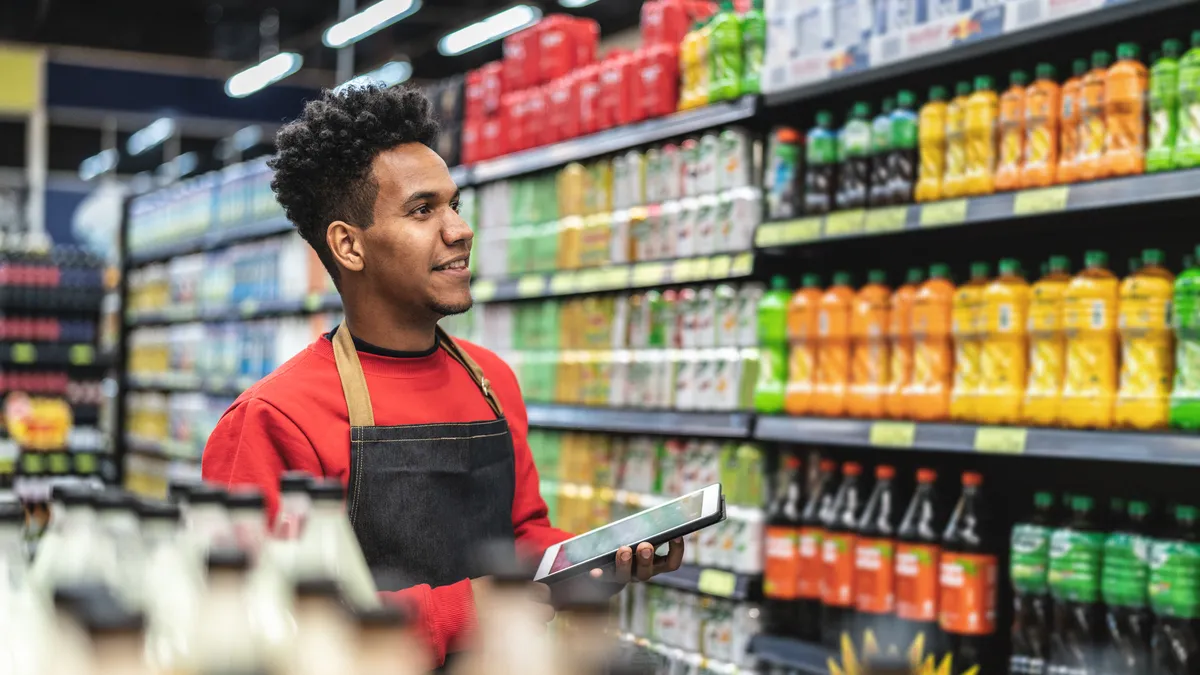 A grocery clerk holding a tablet in a soda aisle at a grocery store.