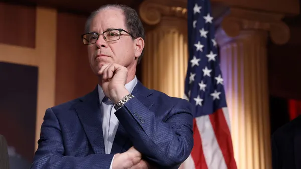 A White man wearing a blue suit and glasses looks out of frame pensively. He is standing in front of an American flag on a flag pole.