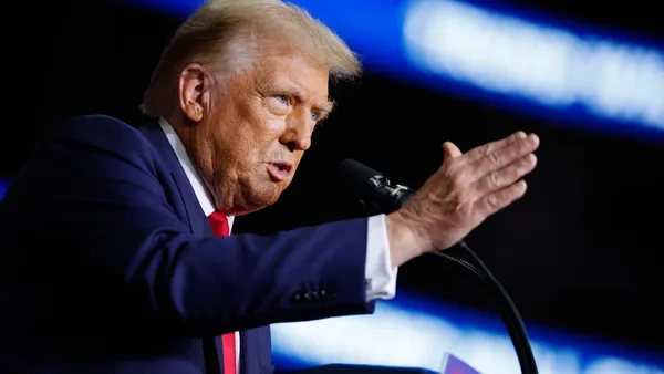 Donald Trump gestures while speaking into a microphone, with blue and white text blurred in the background.