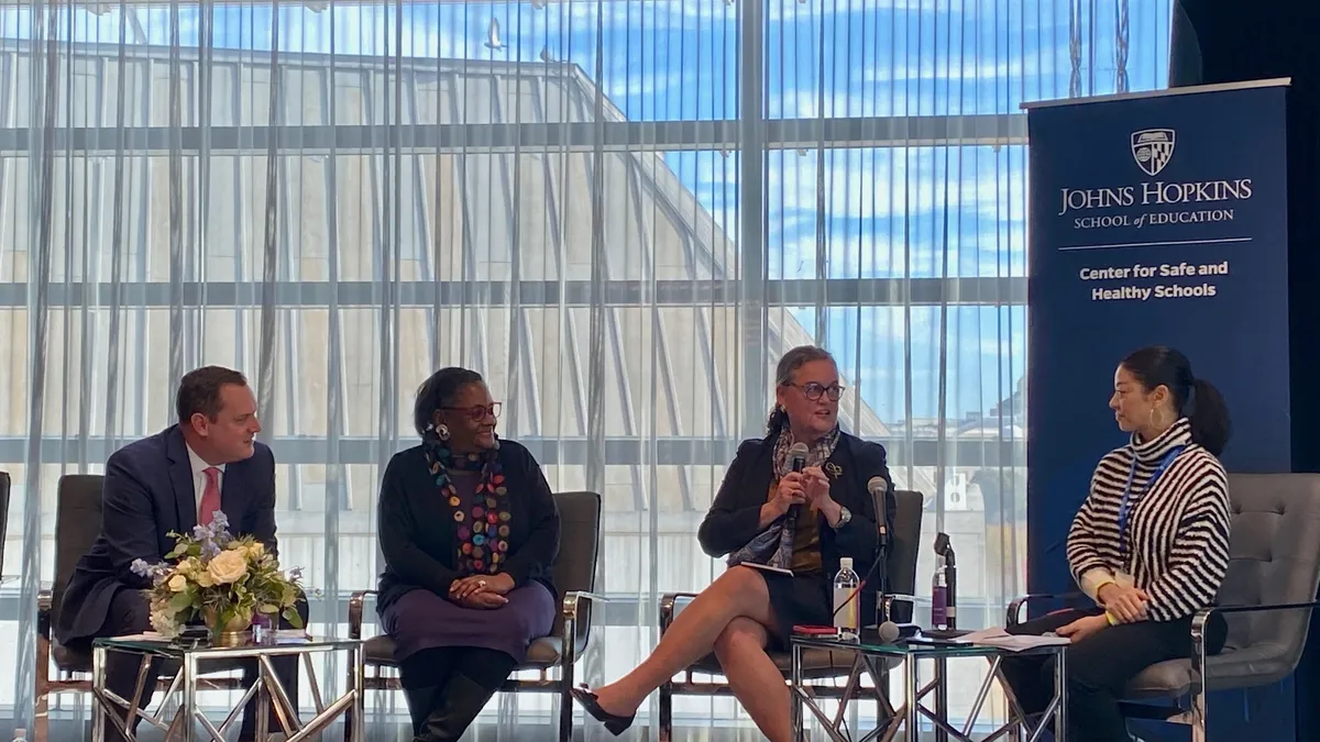 Four adults sit on a stage. One has a microphone. A glass window is behind them.