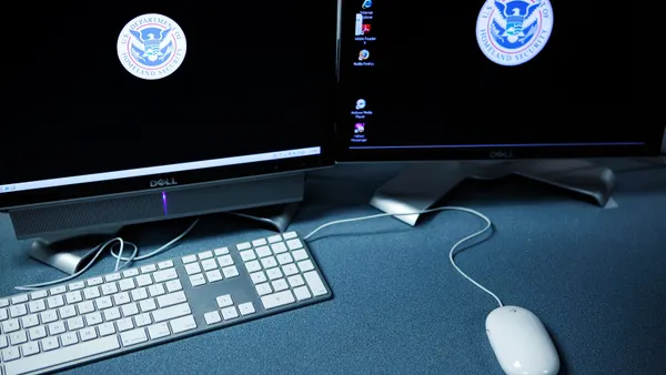A desk holds a mouse, a keyboard, and two computer screens with black wallpaper and the Department of Homeland Security logo