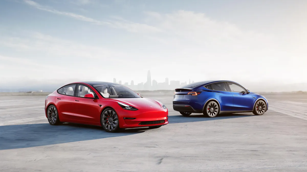 A red Tesla Model 3 and blue Tesla Model Y parked with a city skyline in the background.