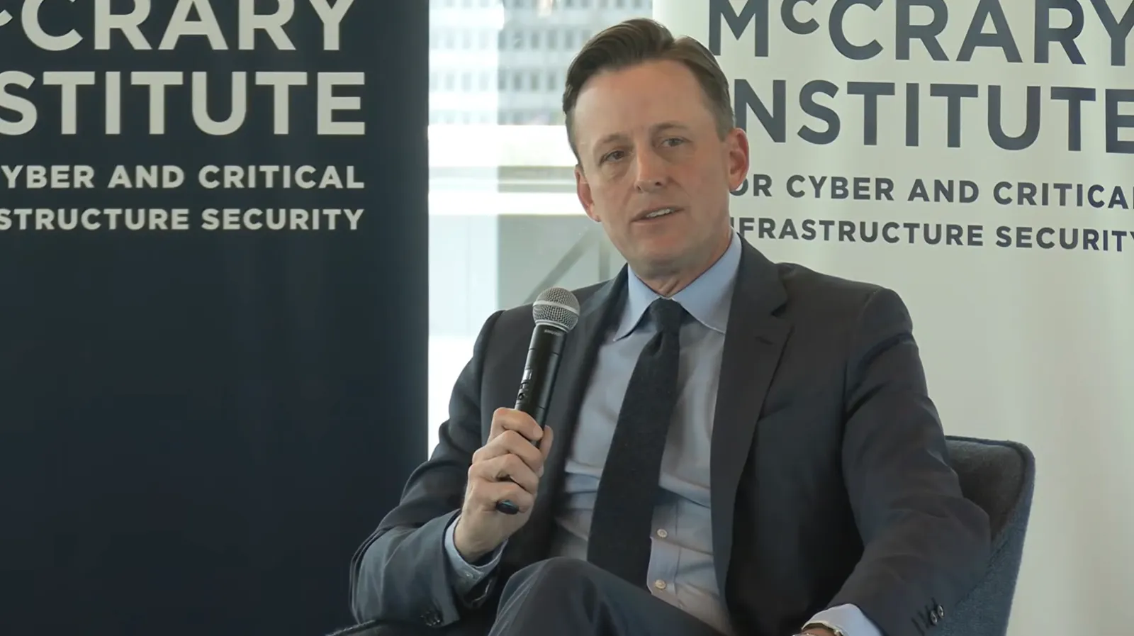 A man holds a microphone while sitting in a chair and looking off-camera