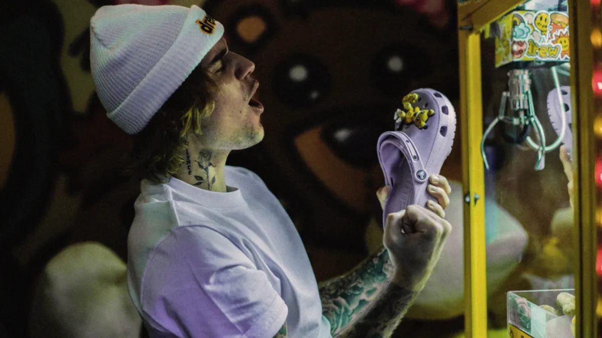 Justin Bieber, wearing a beanie and T-shirt, holds up a Croc shoe in front of a claw machine