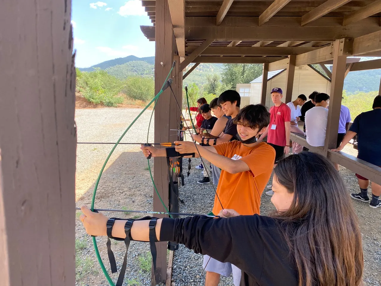 Six students are shown lined up with bows drawn at an archery range as an instructor watches on. Other adults are seated in the background.