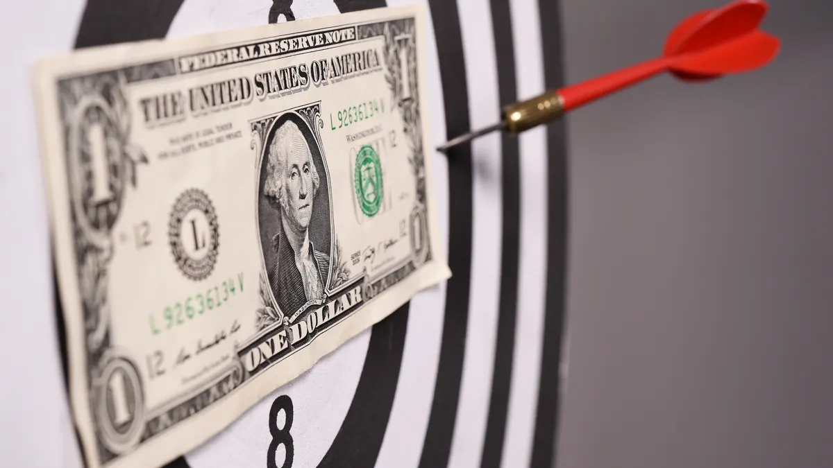 A dart board with a dollar bill at the center, with a dart stuck to it that has missed the bullseye and the dollar bill