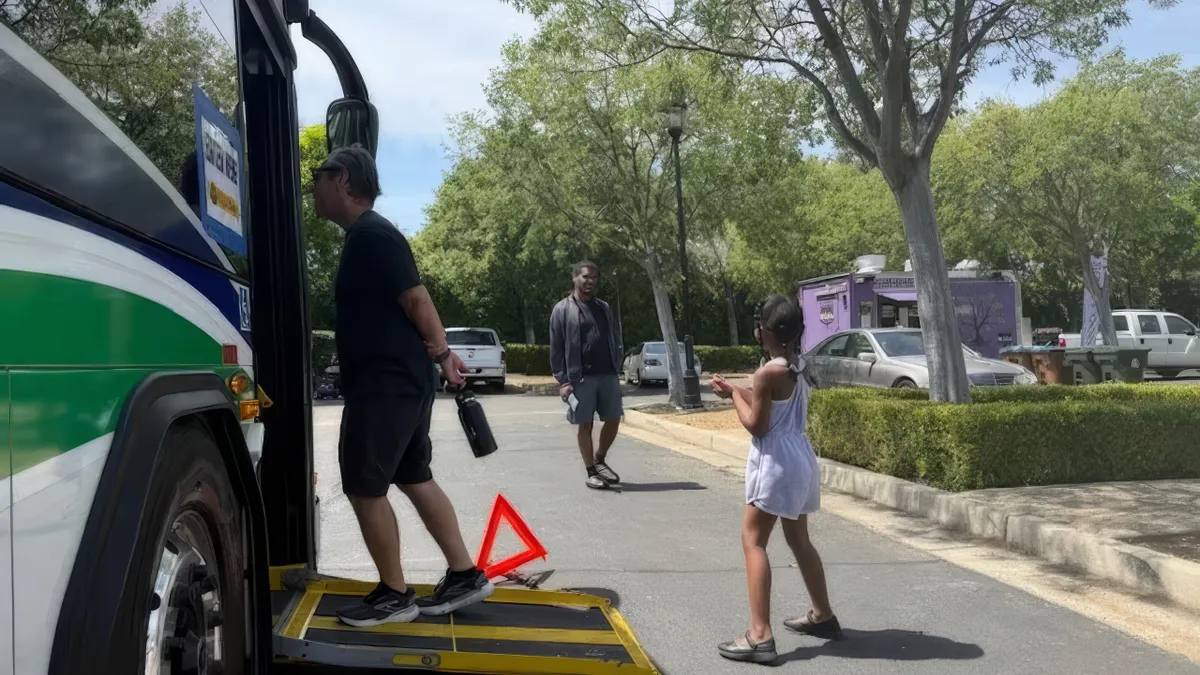 A man walks up a ramp to a transit bus on a tree-covered street with a young girl and another man to the side.