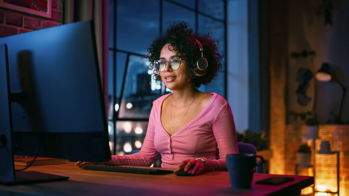 A Black woman wearing headphones uses her computer at home, at night