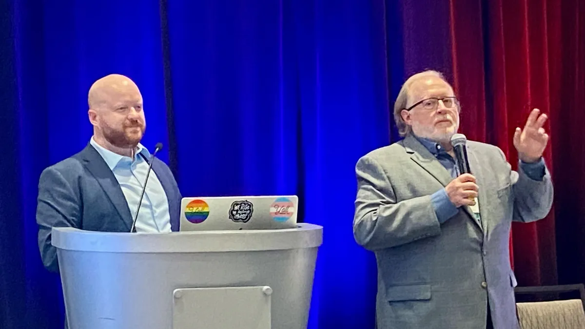 Two adults stand in front of a dark curtain. One is standing behind a podium that has a laptop open. The other adult is standing and holding a microphone near their face.