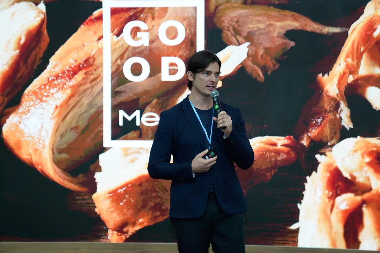 Josh Tetrick speaks into a microphone standing in front of a wall-sized image featuring pieces of Good Meat chicken.