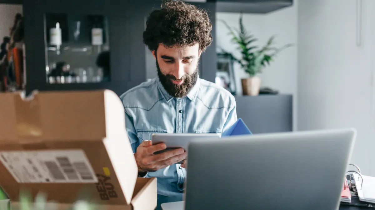 A person sits at a table with a laptop computer and Best Buy product box.