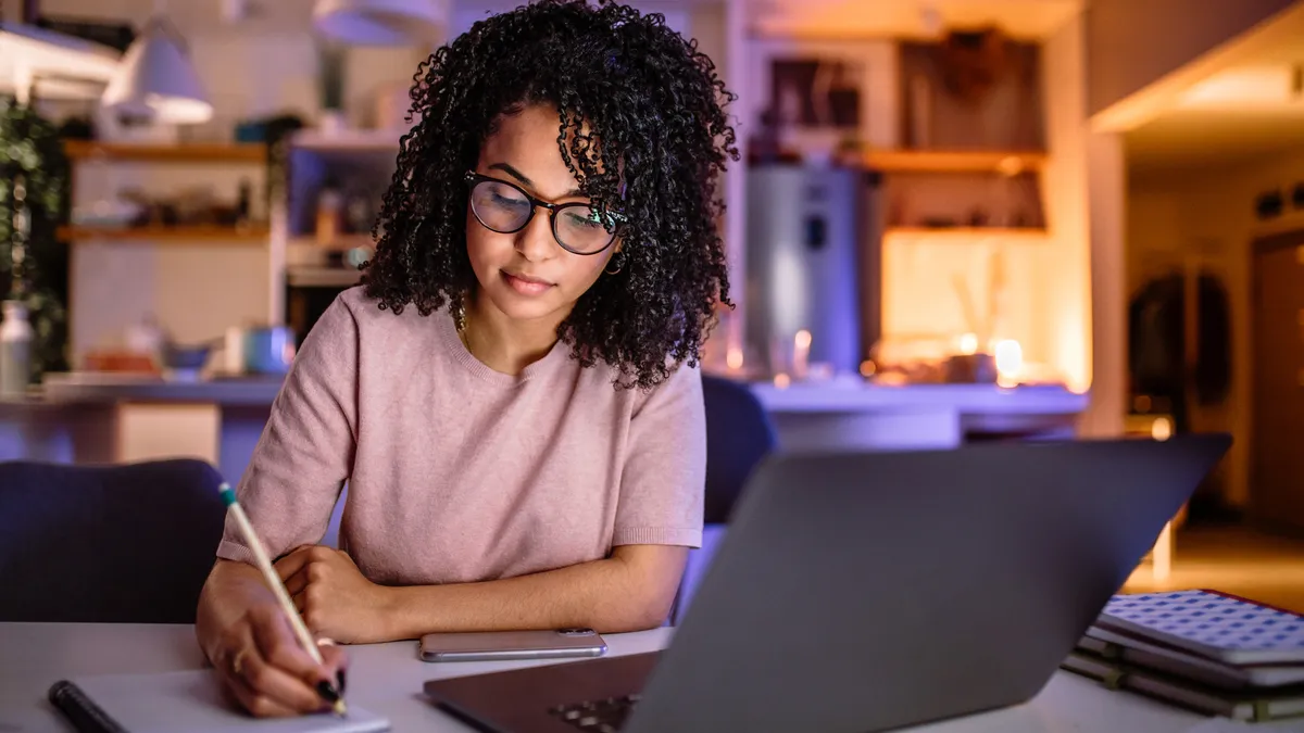 A person writes in a notebook while a laptop sits open on the desk.