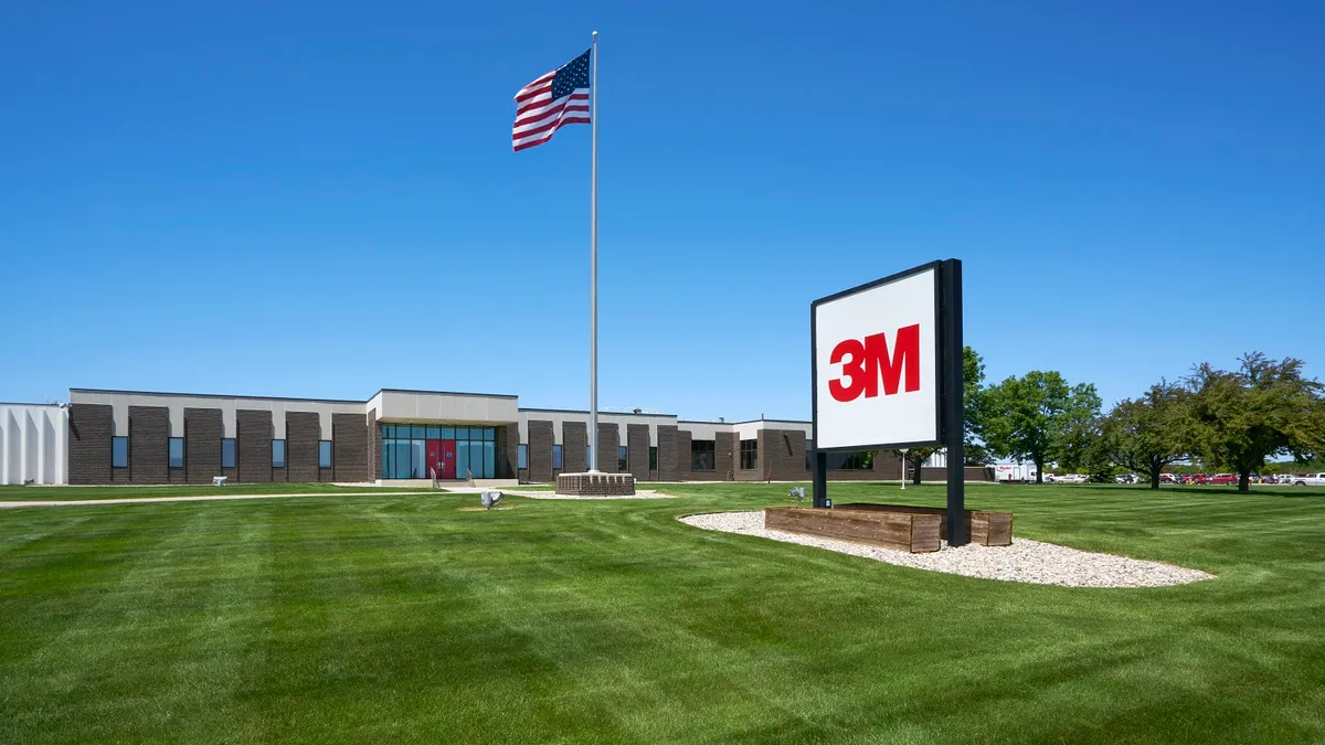 A brown and white building with a green lawn and a red and white 3M sign in front of it on a clear and sunny day.