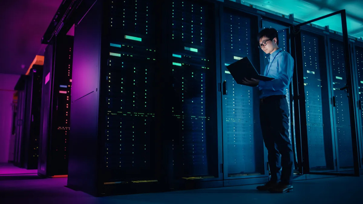 In data center, male IT technician running maintenance program on a laptop for server rack of modernized enterprise computer system.