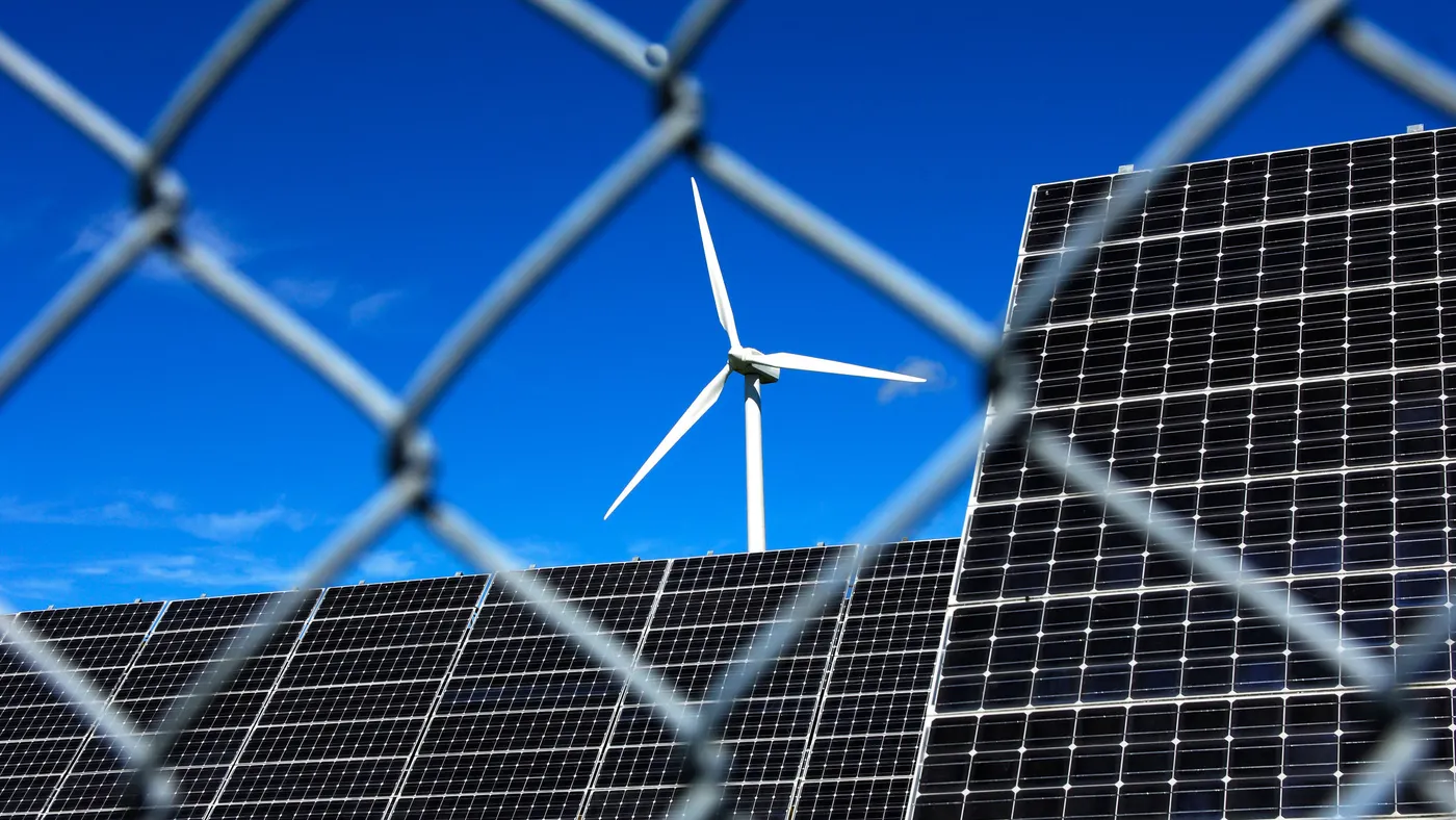 A wind turbine and large solar panel array viewed through a chain-link fence, set against a bright blue sky