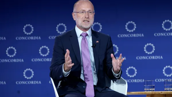 A man wearing a blue suit and purple tie gestures with both hands while sitting in a chair on a stage