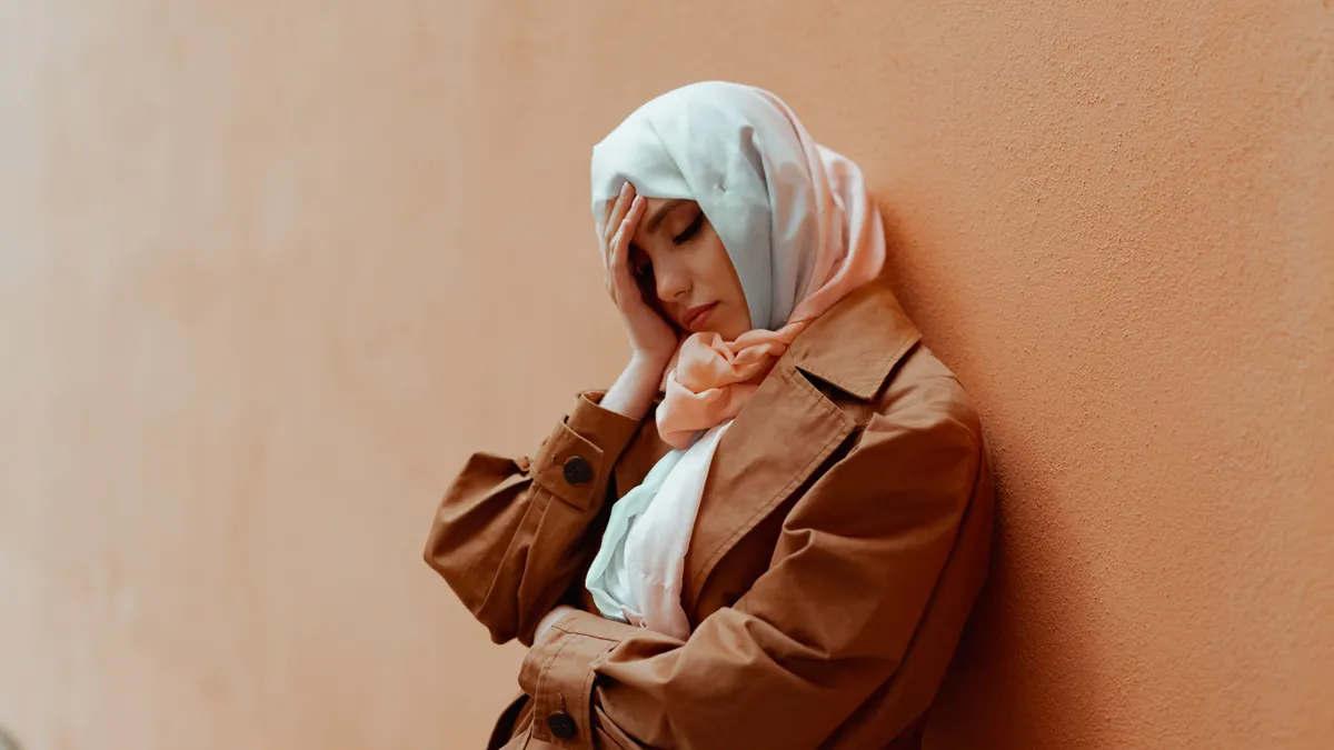 A distressed person with a hijab stands against the wall