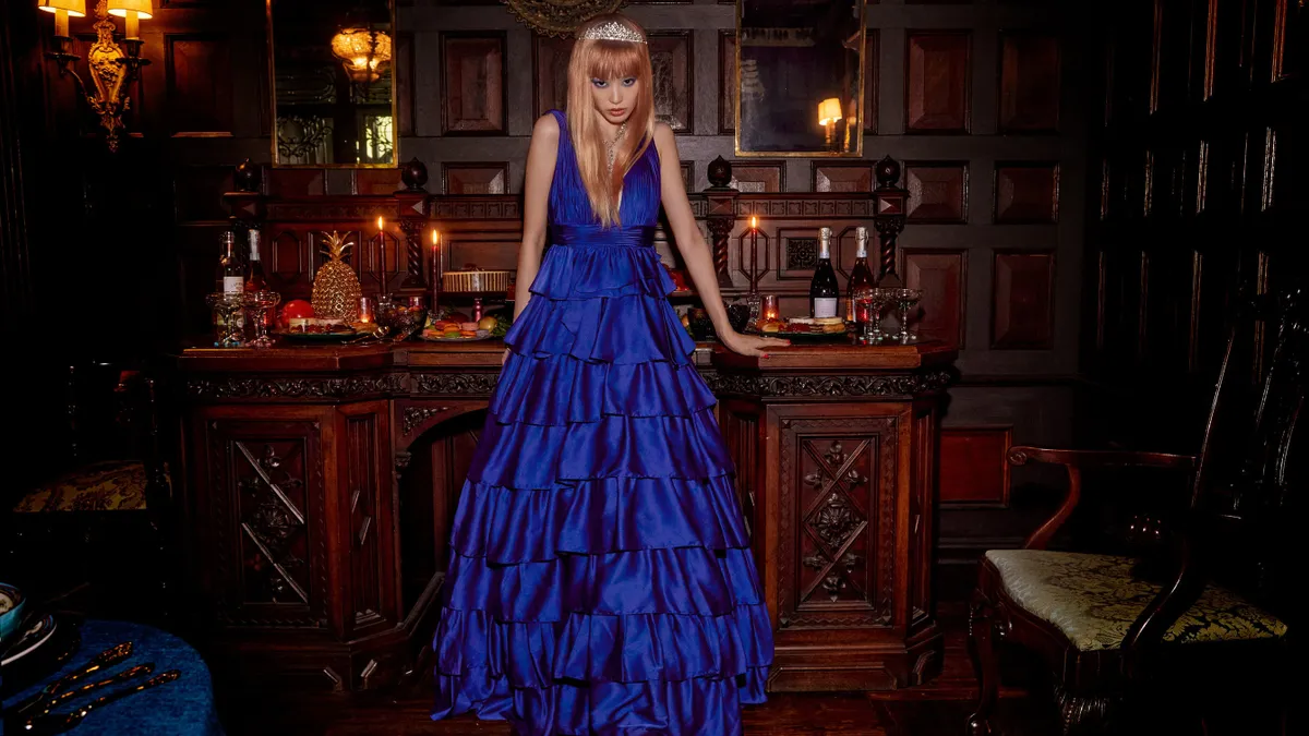 In a dimly lit room, a party-goer in a long blue dress, wearing a tiara, stares into the camera.