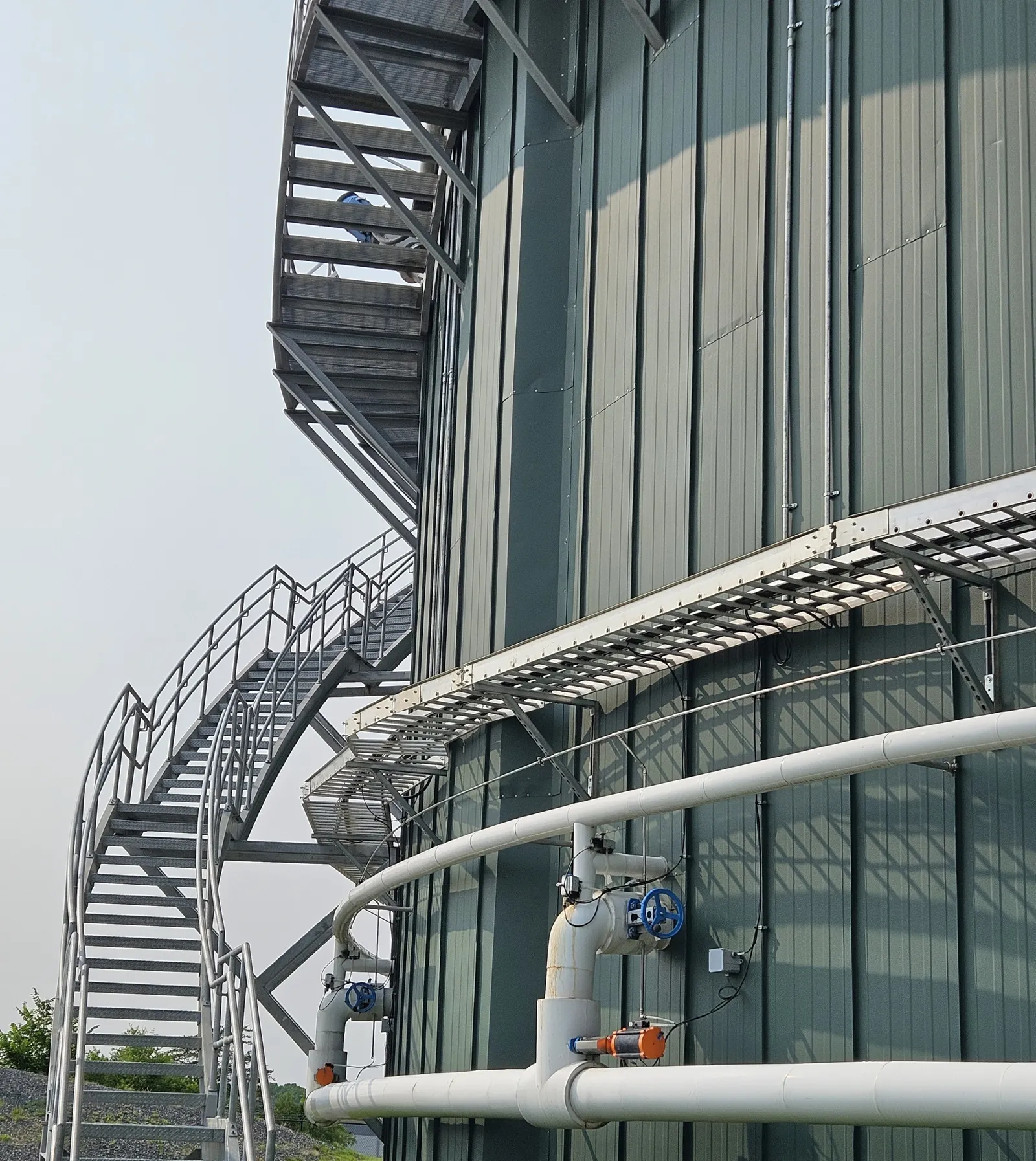 The side of a large cylindrical building with a catwalk snaking up the exterior.