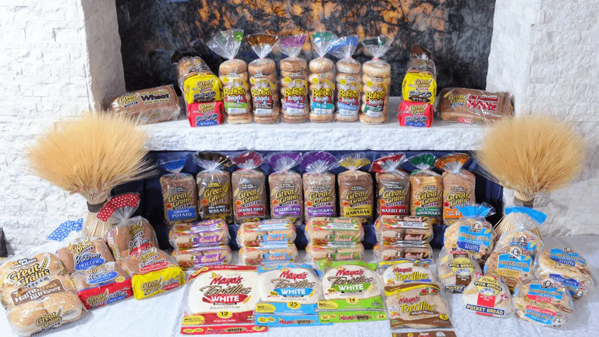 Packages of Papa Pita Bakery's buns, bread, bagels, English muffins,tortillas and flatbreads arranged on a white brick shelf area with wheat sheafs in the background.