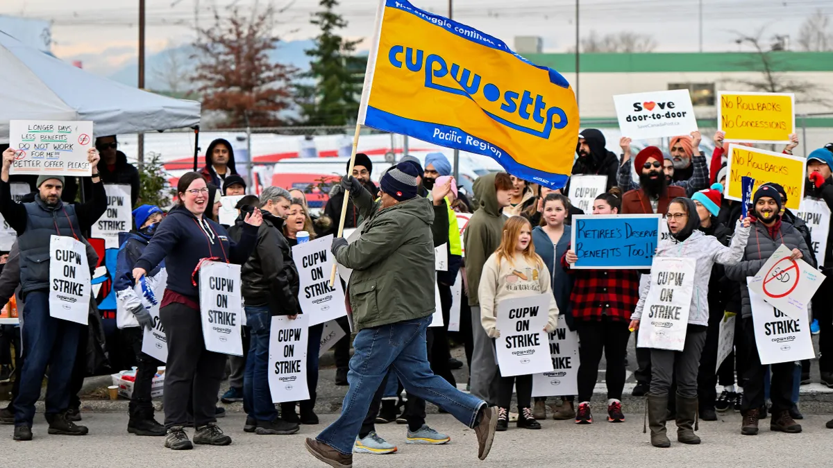 A person runs while holding a Canadian Union of Postal Workers flag in front of a picket line.