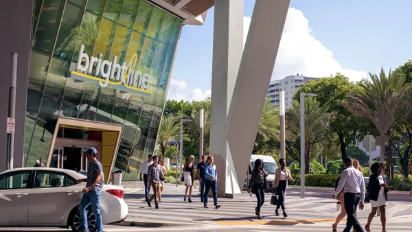 People walking on a plaza to and from a glass-front building with a sign reading "Brightline."
