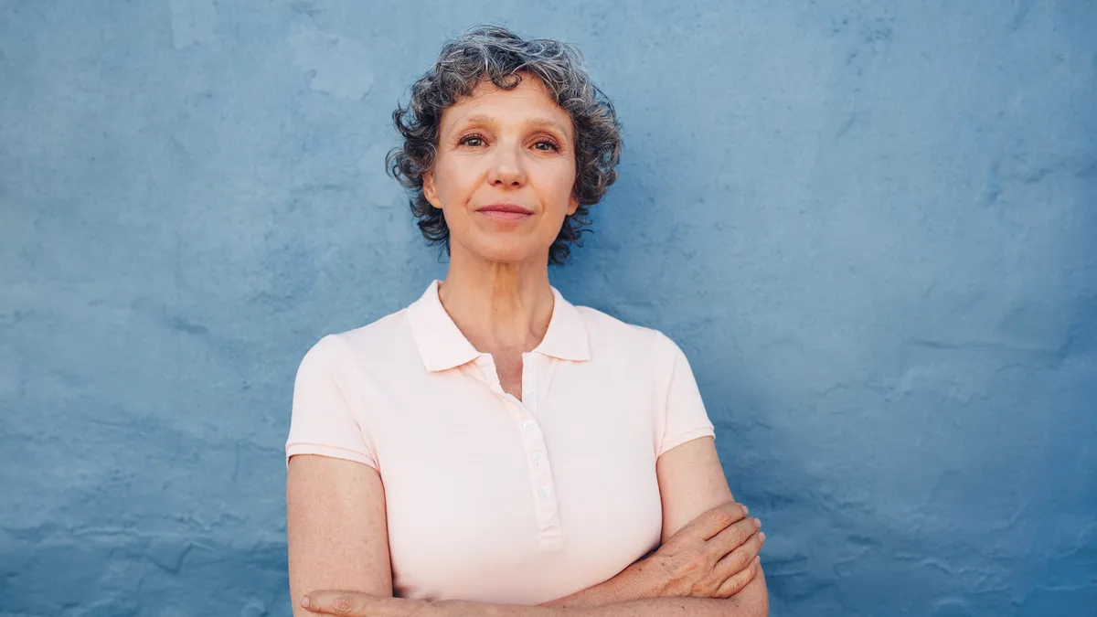 Confident "mature" person standing in front of a soft blue wall with their arms crossed