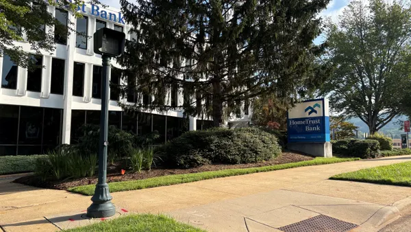 A sign in front of a white and black office building and next to an evergreen tree reads "HomeTrust Bank."