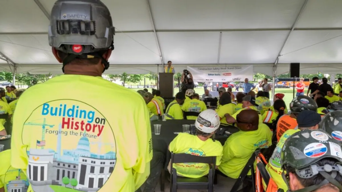 People in bright yellow shirts and hard hats gather under a tent at an event.