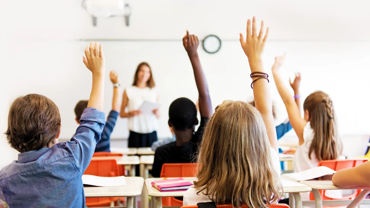 Students with raised hands sit at desks facing a teacher at the front of a classroom.