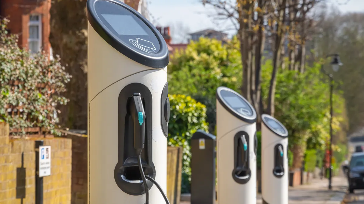 A row of white EV chargers with cables and plus stand against a blurry background of trees and a brick building.