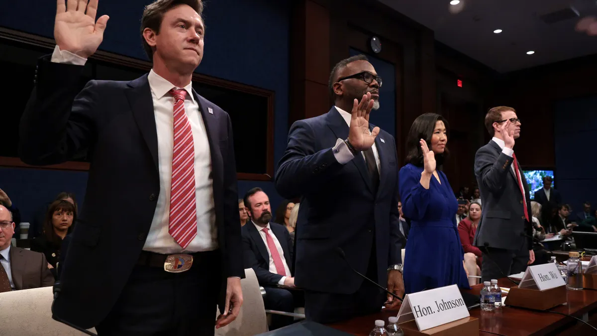 Three people wearing dark suits and one person wearing a blue dress stand with their hands raised.