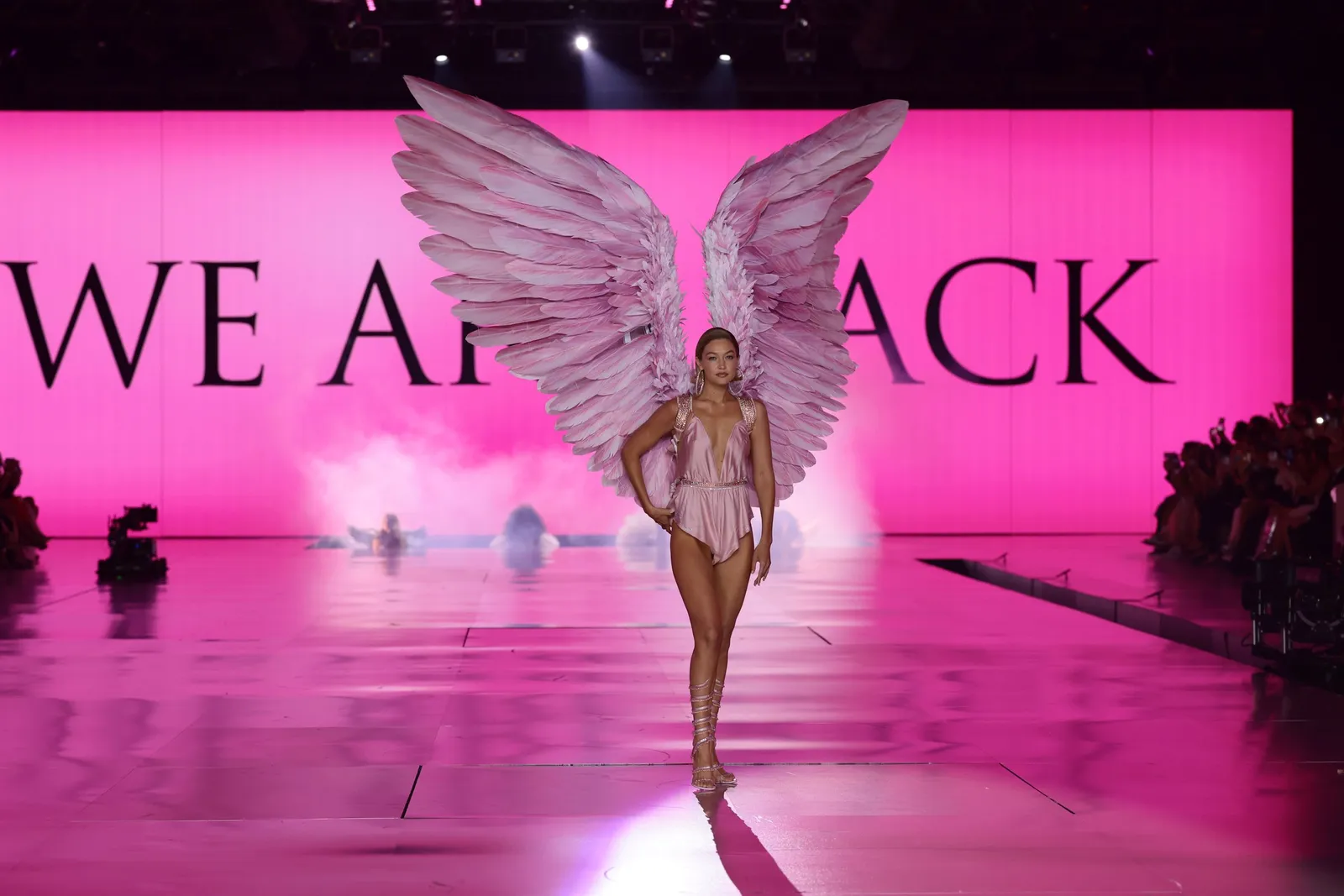 A model wearing pink with wings stands in front of a sign reading &quot;we are back&quot;