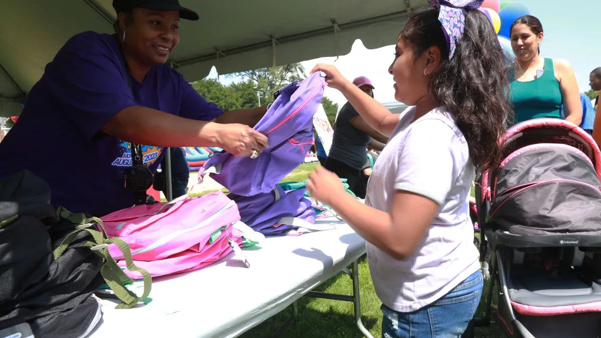 A student receives a backpack from an adult while they are outside. The adult is under a tent and behind a table. In the background is another adult with a stroller.