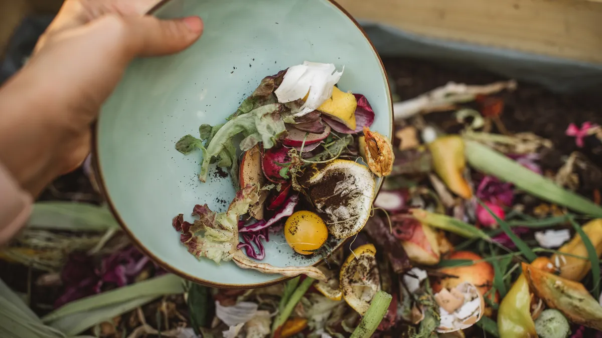 A hand brushes organic materials like vegetable scraps off a plate and into a bin