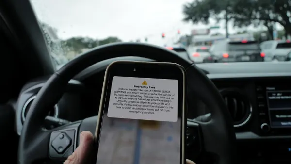 A phone showing a severe weather alert in front of a steering wheel with traffic out the front window.