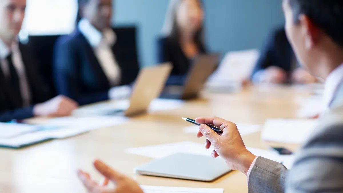 A businessman gestures with a pen in hand  while seated at a table with other stakeholders in aconference room.