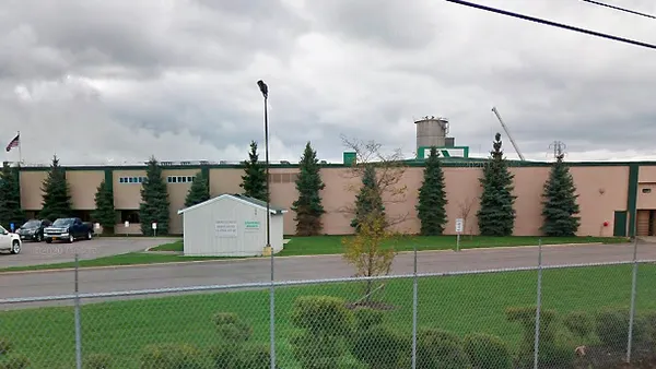 The exterior of an industrial facility with cars in a parking lot, which is surrounded by a chain-link fence.