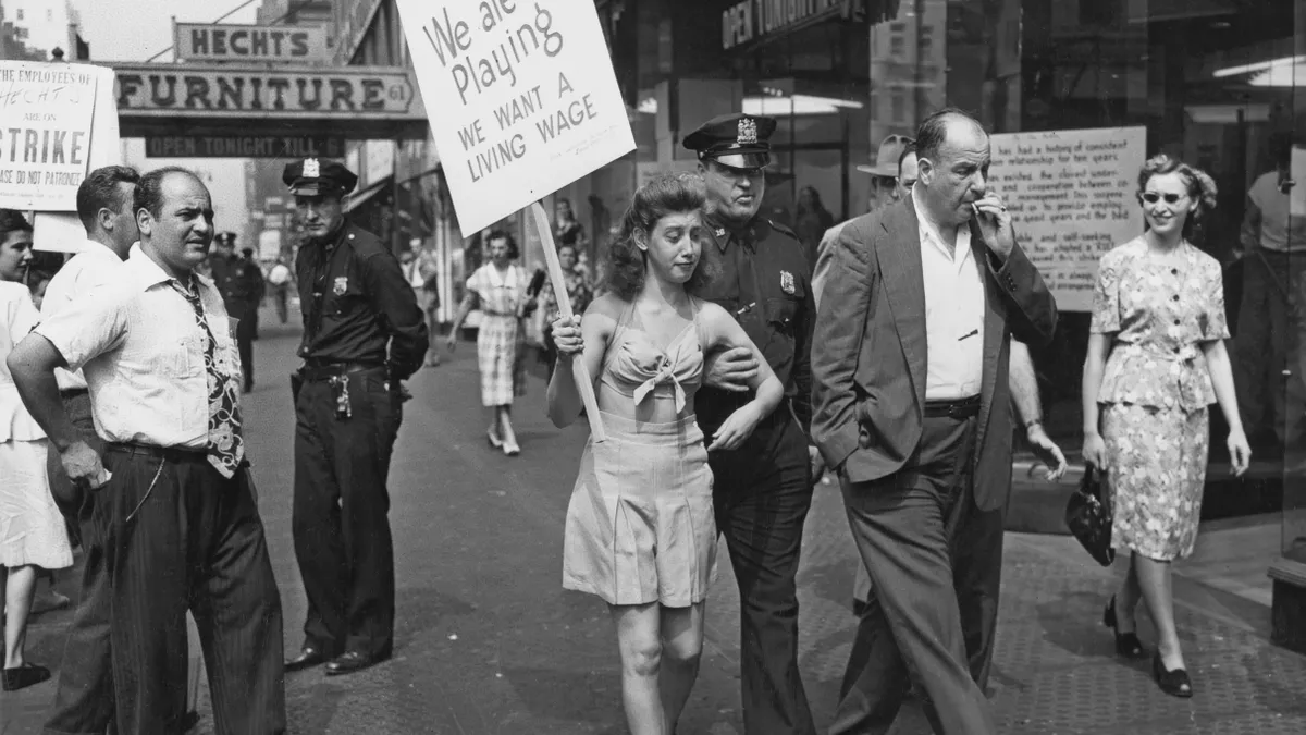 Passersby stand and watch as a police officer escorts a demonstrator holding a placard reading 'Hecht's employees on strike, we are not playing, we want a living wage'