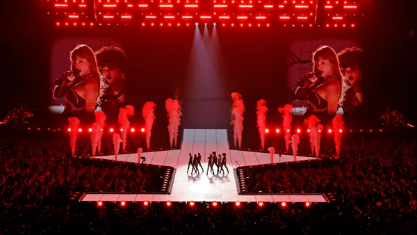 Taylor Swift performing live onstage flanked by dancers in front of a stadium crowd. In the background are bursts of smoke and large screens featuring images of Swift.