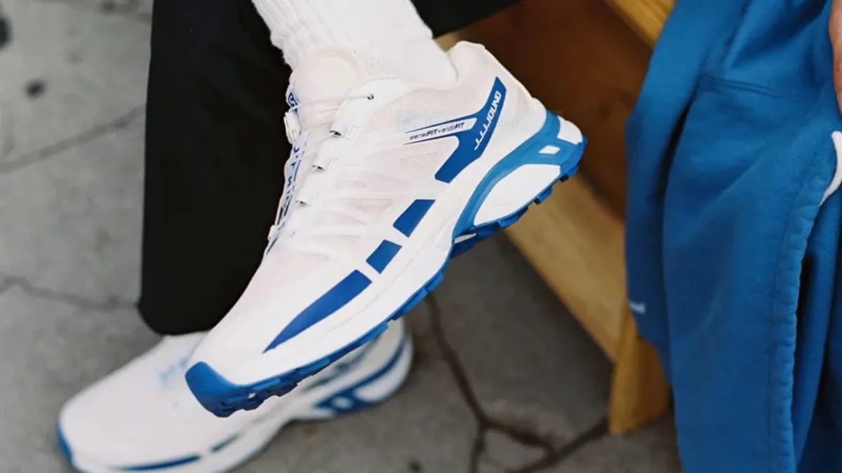 A picture of a person's foot wearing a white and blue sneaker by Salomon and JJJJound.