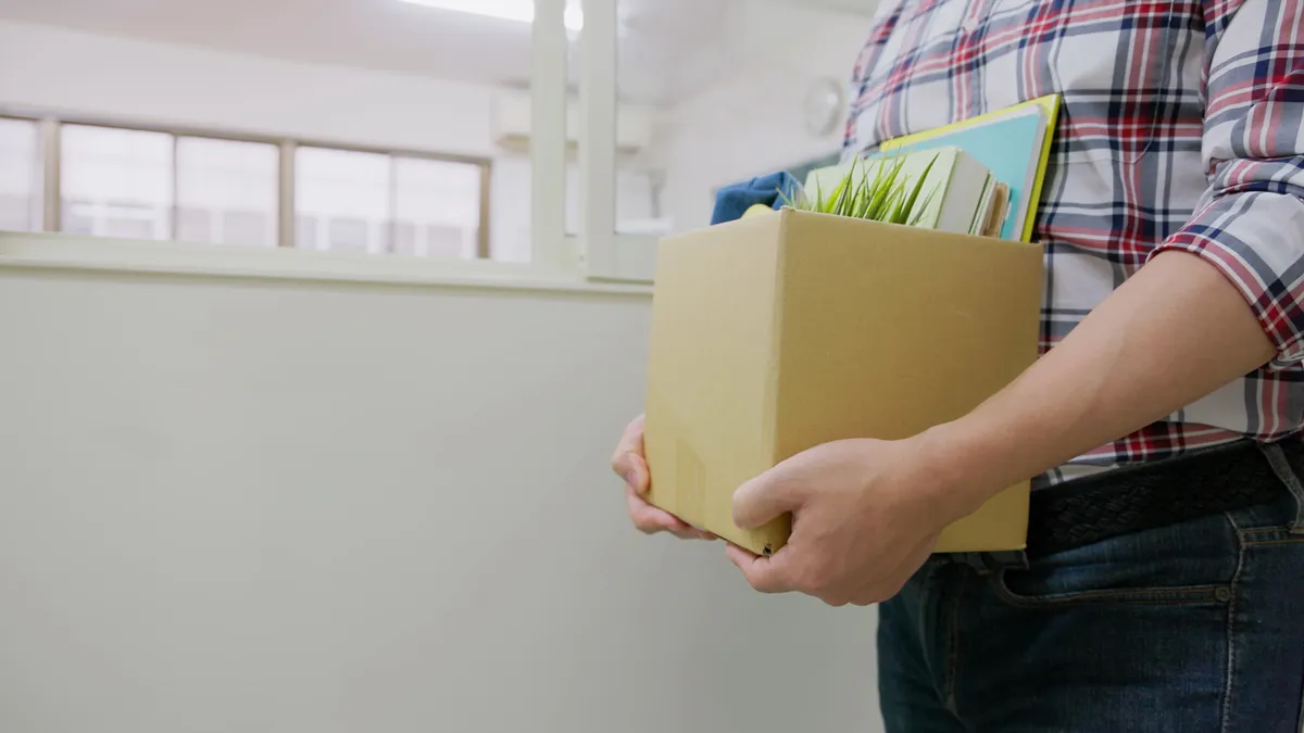 An adult holds a box full of office supplies on their way out of a classroom.