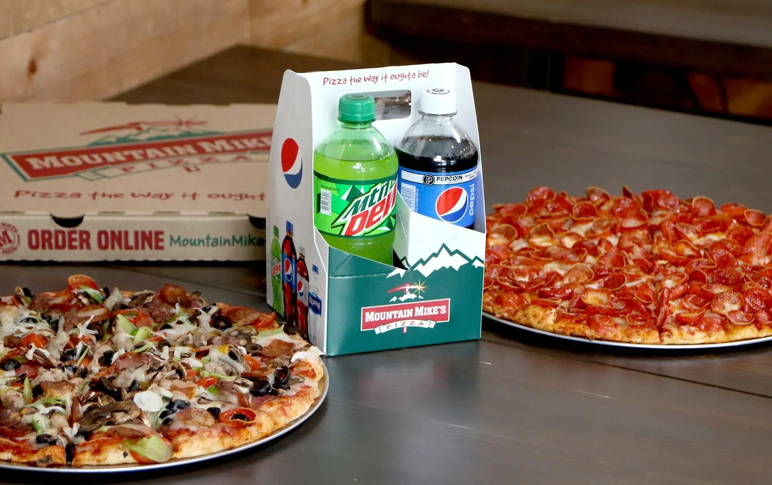 An image of two pizzas on a table with sodas in the center
