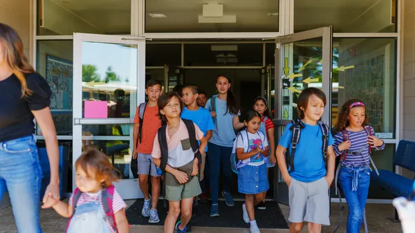 A multiracial group of elementary students of varying ages holding backpacks exit their school through glass doors t the end of the day