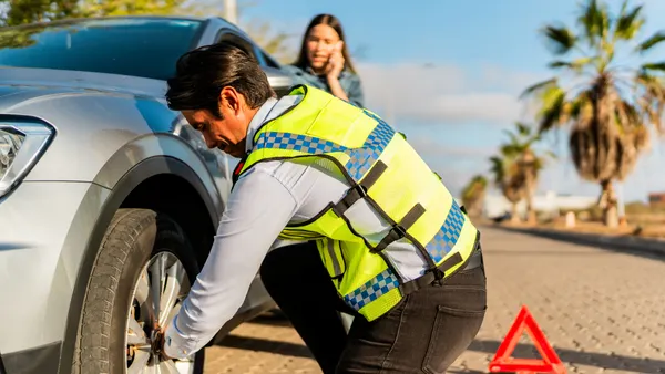 A man changes a tire, while a woman talks on the phone in the background.