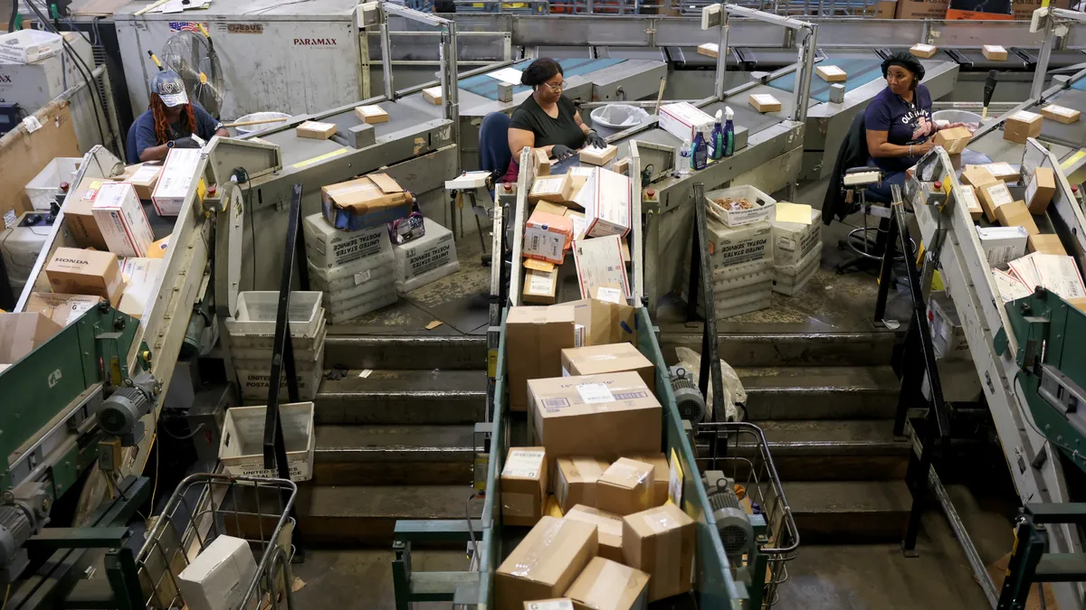 Three U.S. Post Service workers sort packages at a distribution center.