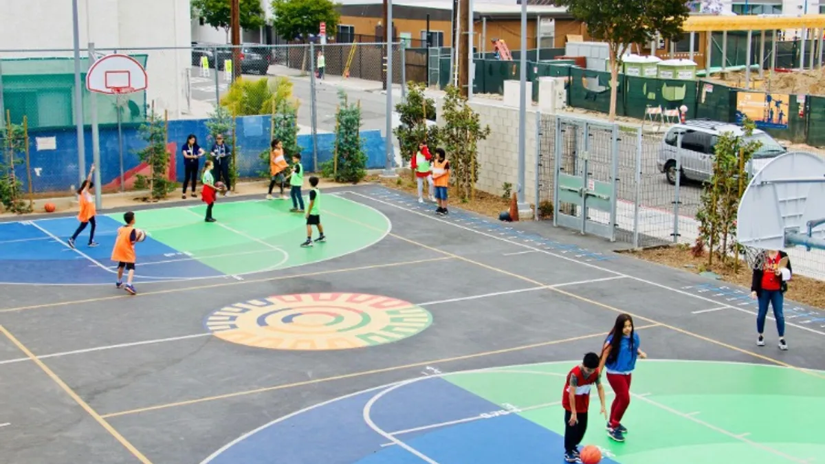 People are moving on an outdoor basketball court. The court has colorful graphics with numbers on each side of the court.