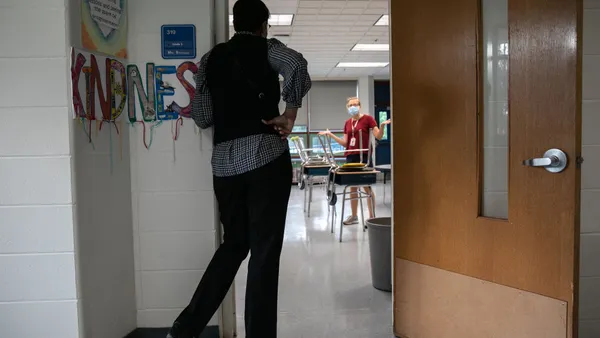 A person stands in the doorway of a classroom with their back to the camera. Another person is seen in the classroom.